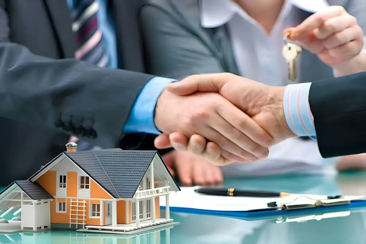 Two people shaking hands over a model house and a set of keys